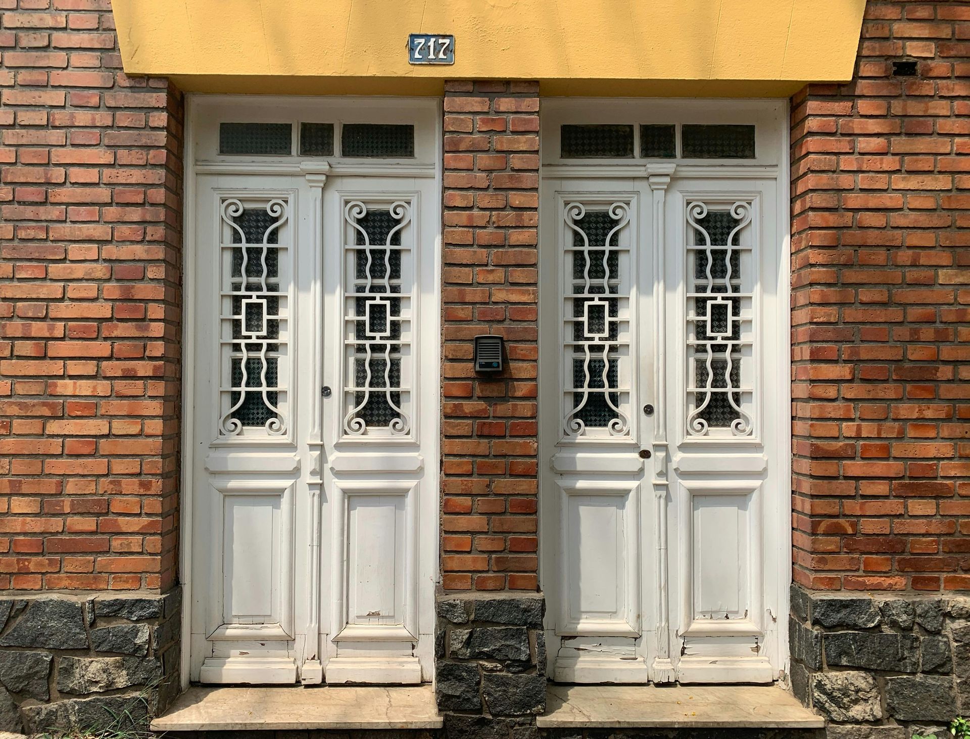 Two white doors with ornate grates set in a brick wall, below a yellow overhang.
