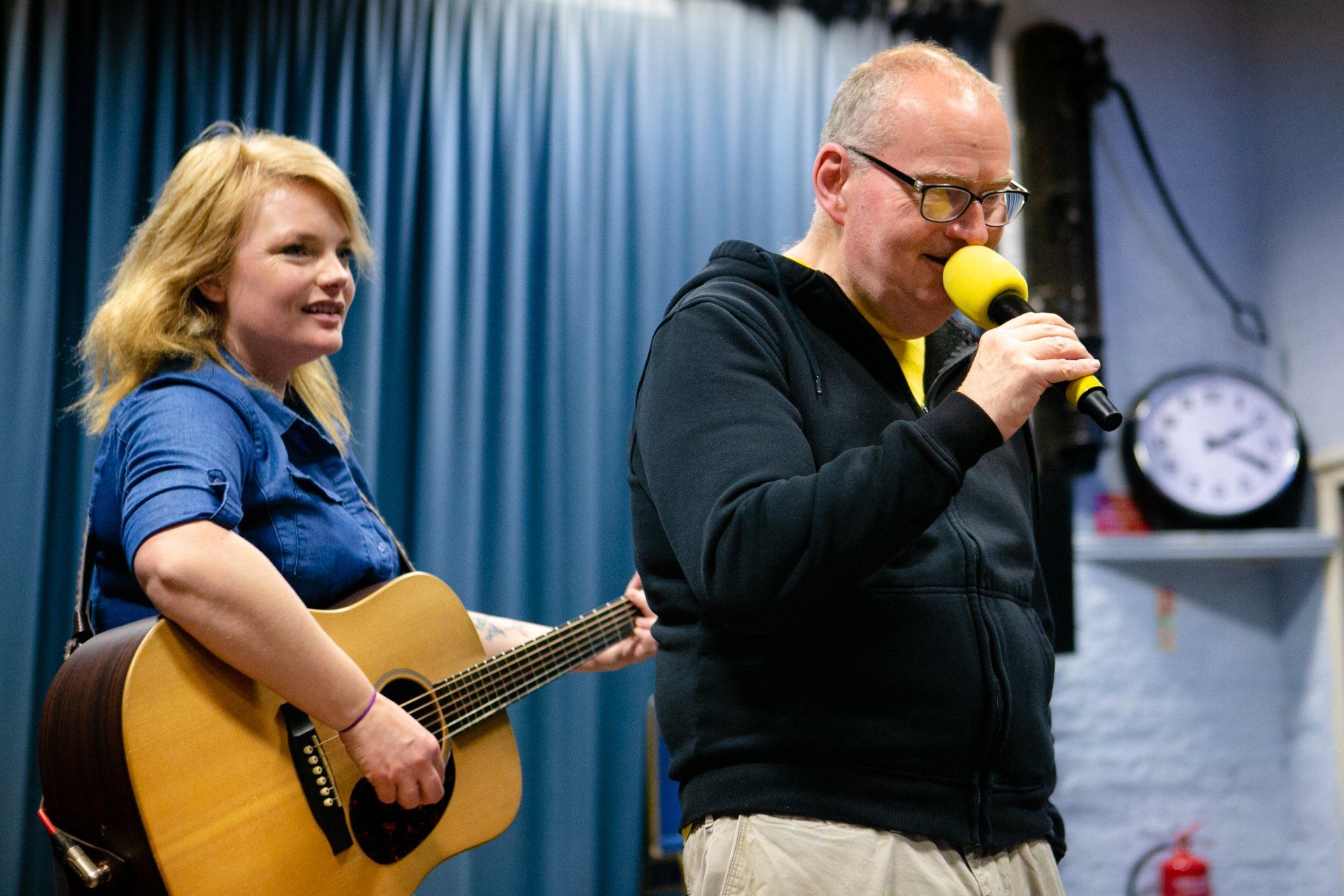 A learning disabled man is holding a microphone and singing. A woman on guitar is supporting his performance.