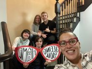 Group posing on stairs with red heart signs reading “I am” and “feel loved,” smiling at the camera.
