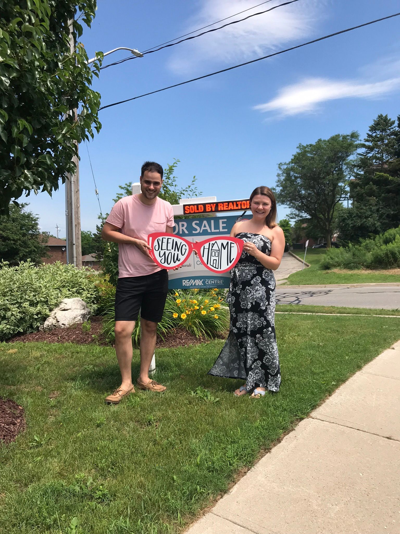 Two people hold a “For Sale” sign on a grassy roadside under a bright blue sky.