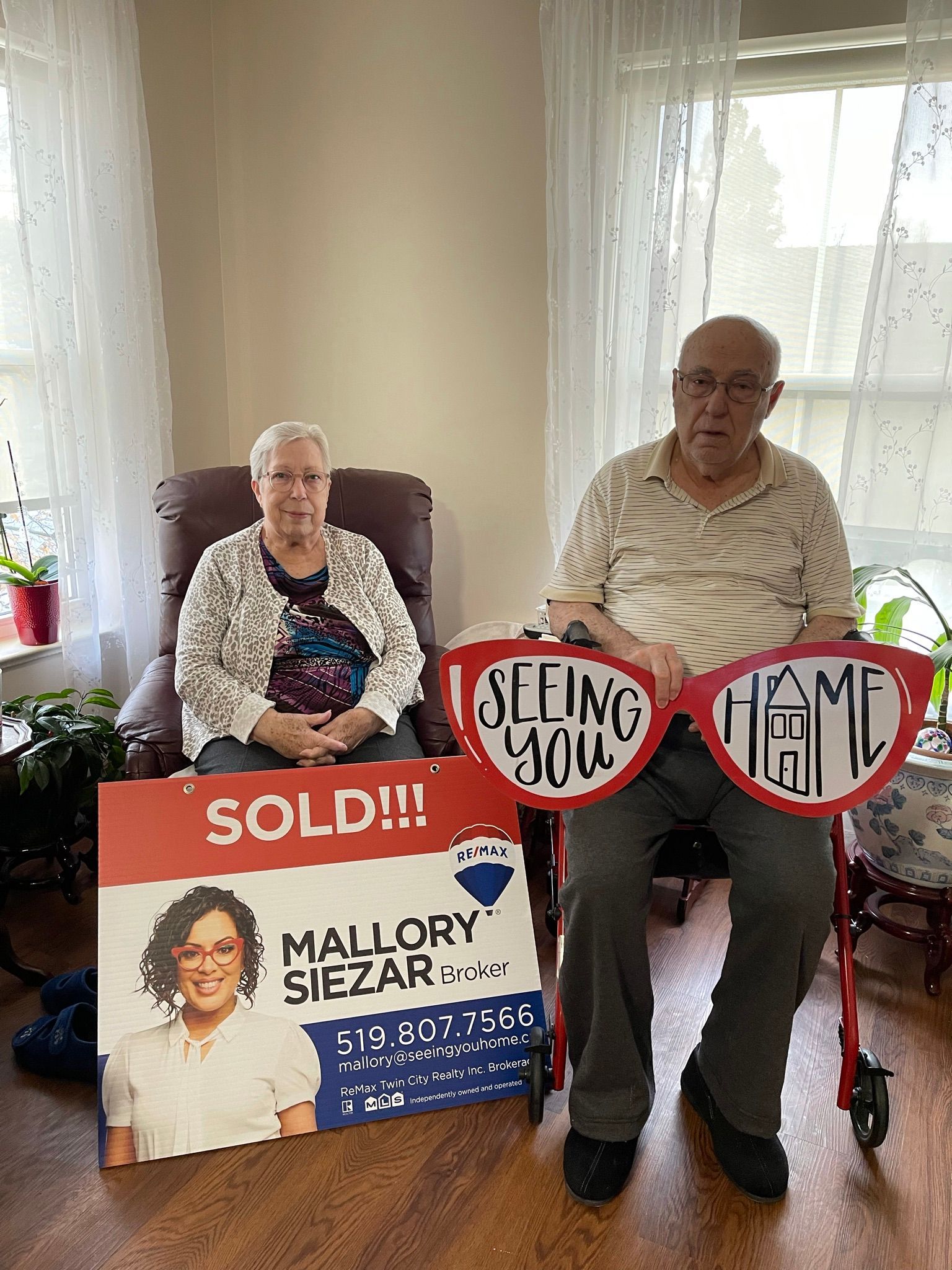 Two seated people hold campaign signs reading “Sold!” and “Keep Them Home” in a bright room
