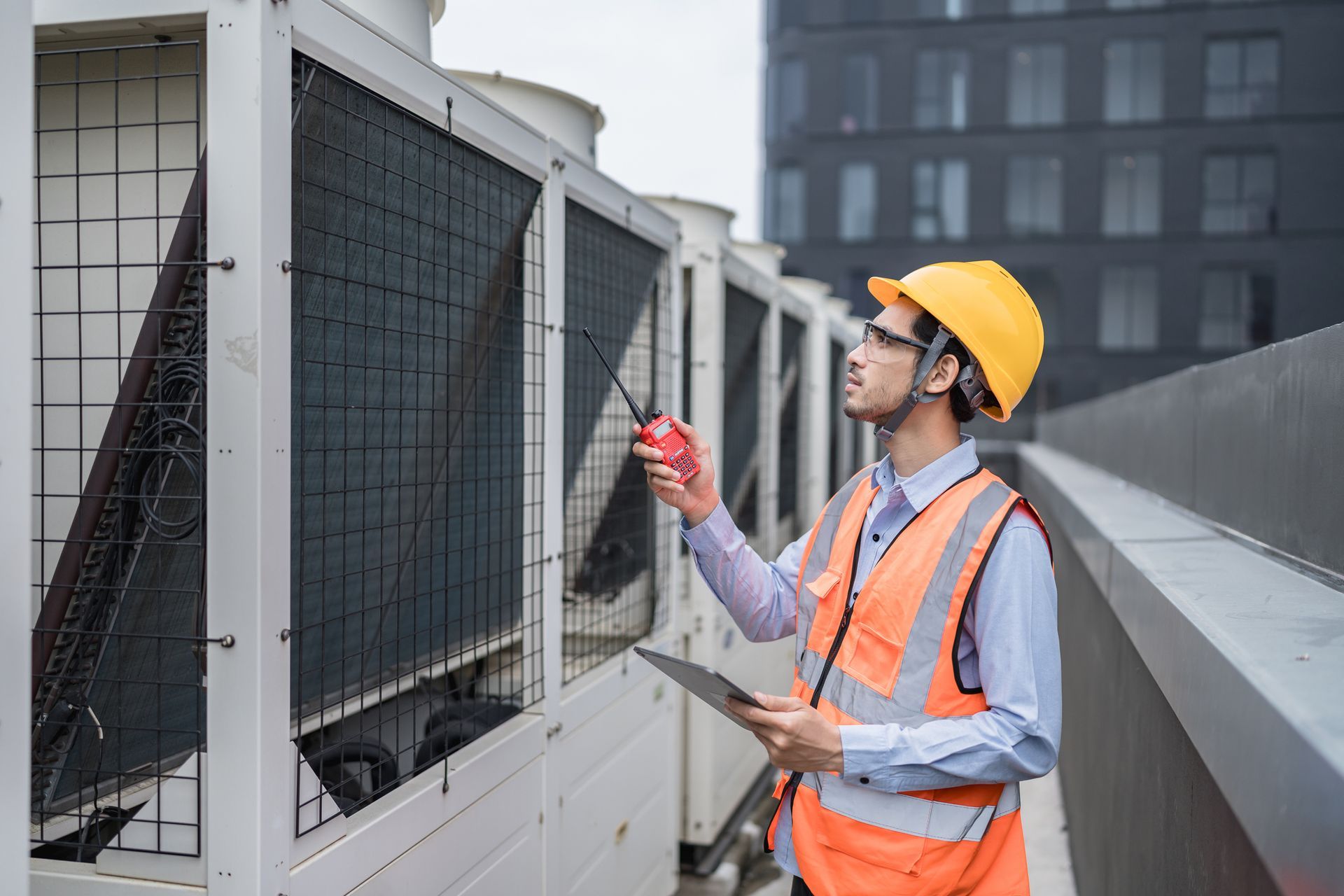 Man in hardhat and vest inspecting HVAC units on a building rooftop, holding a tablet and radio.