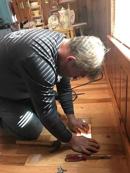 Man kneels on hardwood floor, installing a metal grate; hammer and screwdriver nearby.