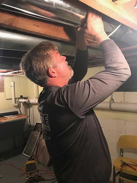 Man working on pipes in a basement. He is wearing a gray long-sleeved shirt and is reaching upwards.