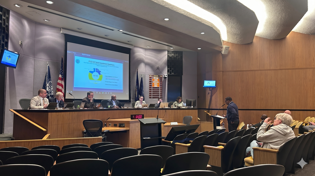 Conference room with a presentation on a large screen, officials at the dais, and a few attendees seated nearby.