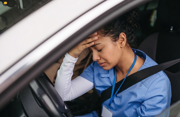 Driver in blue shirt with hand on forehead, looking stressed inside a car with seat belt on.