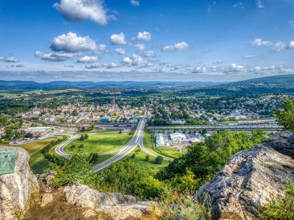 Panoramic view from rocky hilltop over a green valley, town, and winding roads under a bright blue sky.