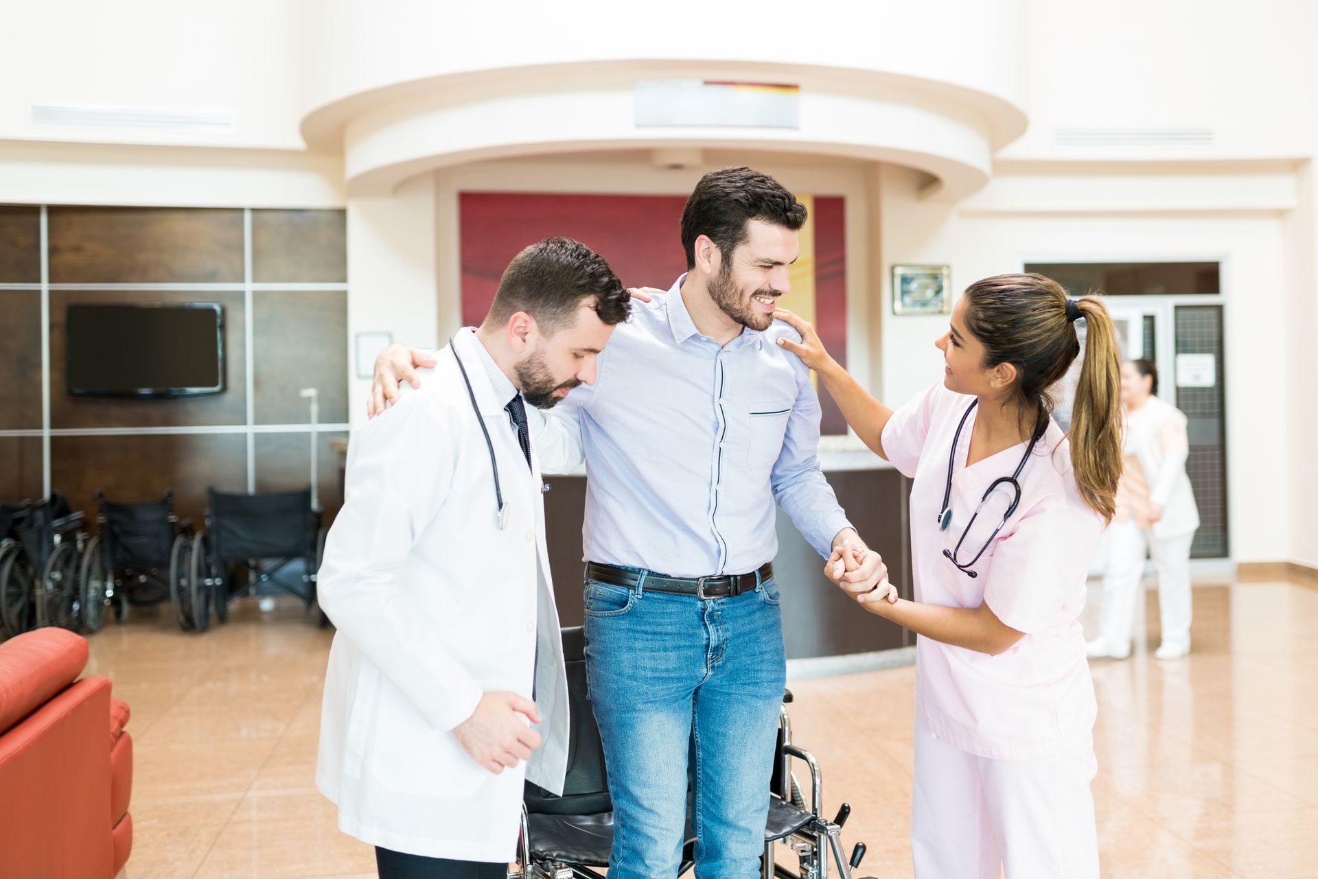 A medical professional and a nurse assist a man in standing up from a wheelchair in a brightly lit hospital lobby.