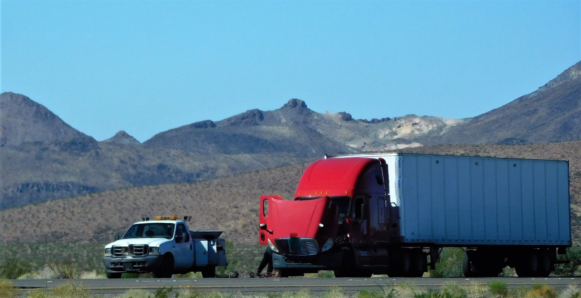A red semi-truck with its hood open is parked on a desert roadside next to a white service truck.