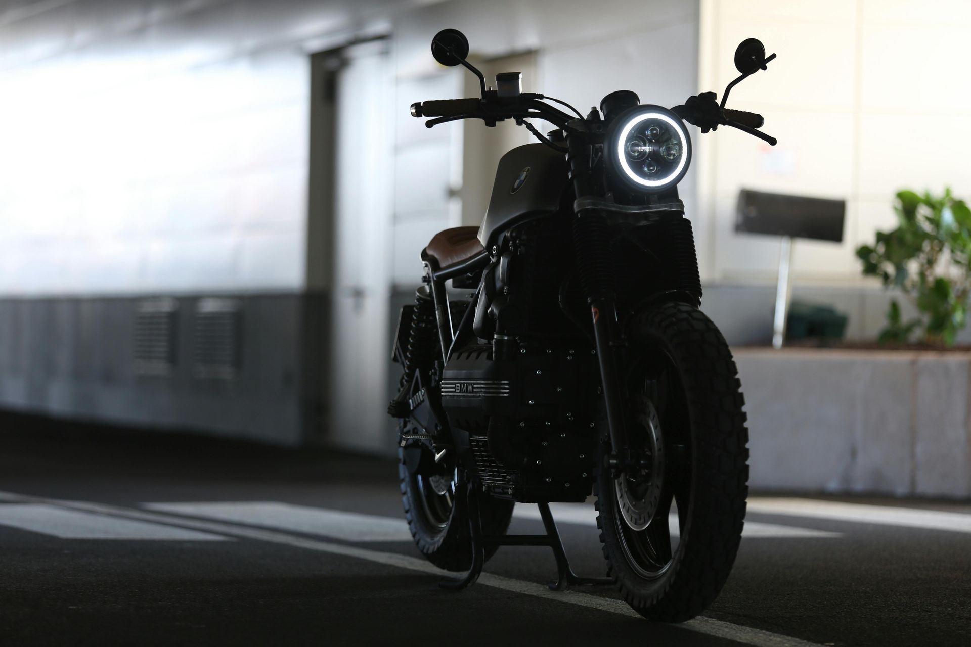 A dark motorcycle with a glowing circular headlight stands parked on a paved surface inside a dimly lit structure.