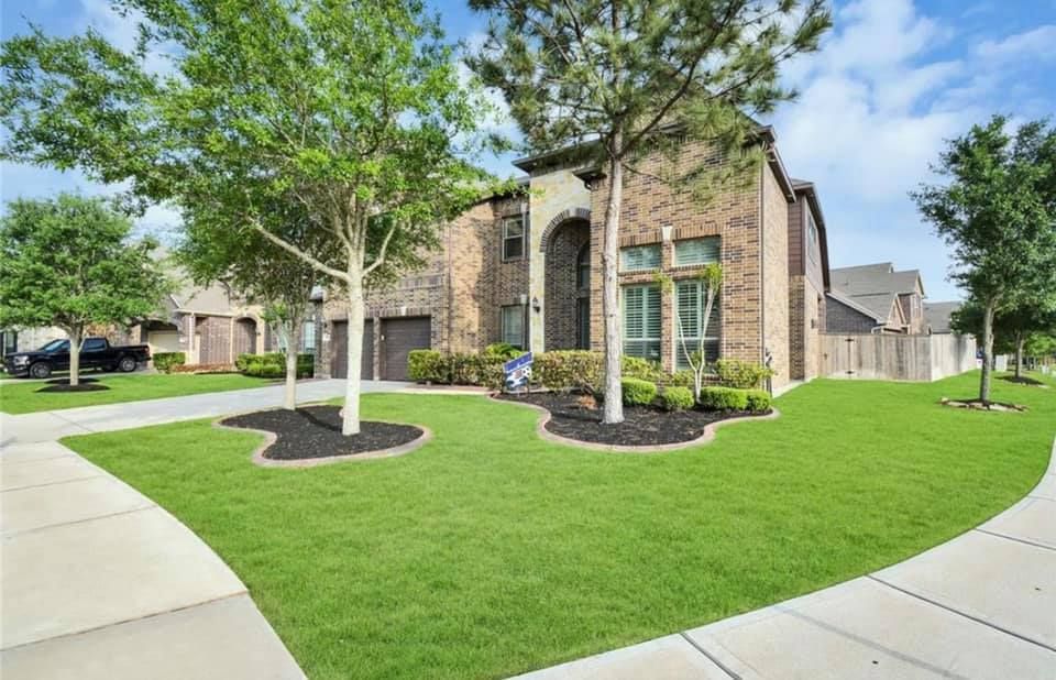 House with a manicured lawn, trees, and driveway. Brick exterior with arched entryway and garage.