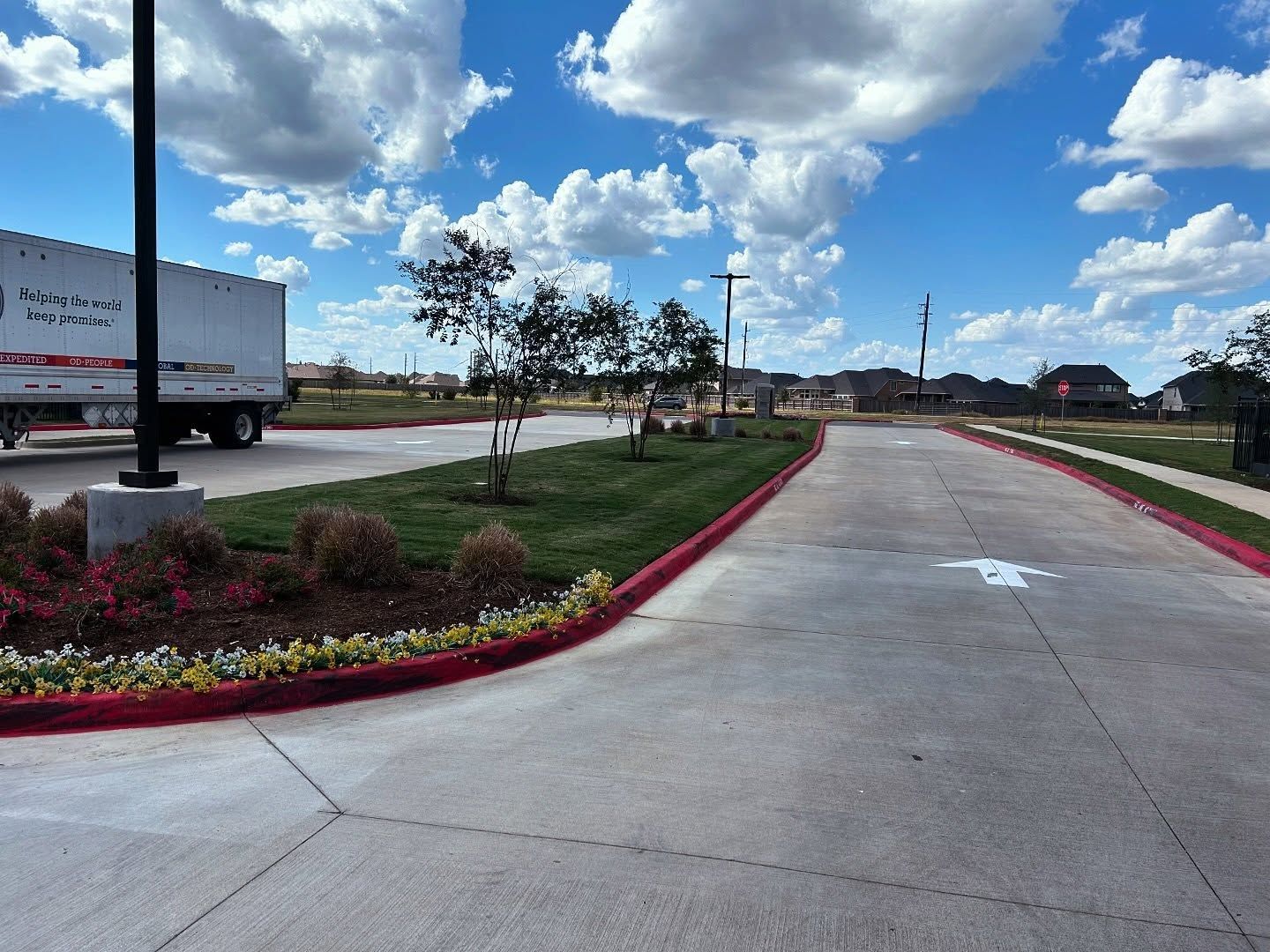 Asphalt road curves past grassy landscaping; a semi-truck sits in the background under a blue sky with clouds.