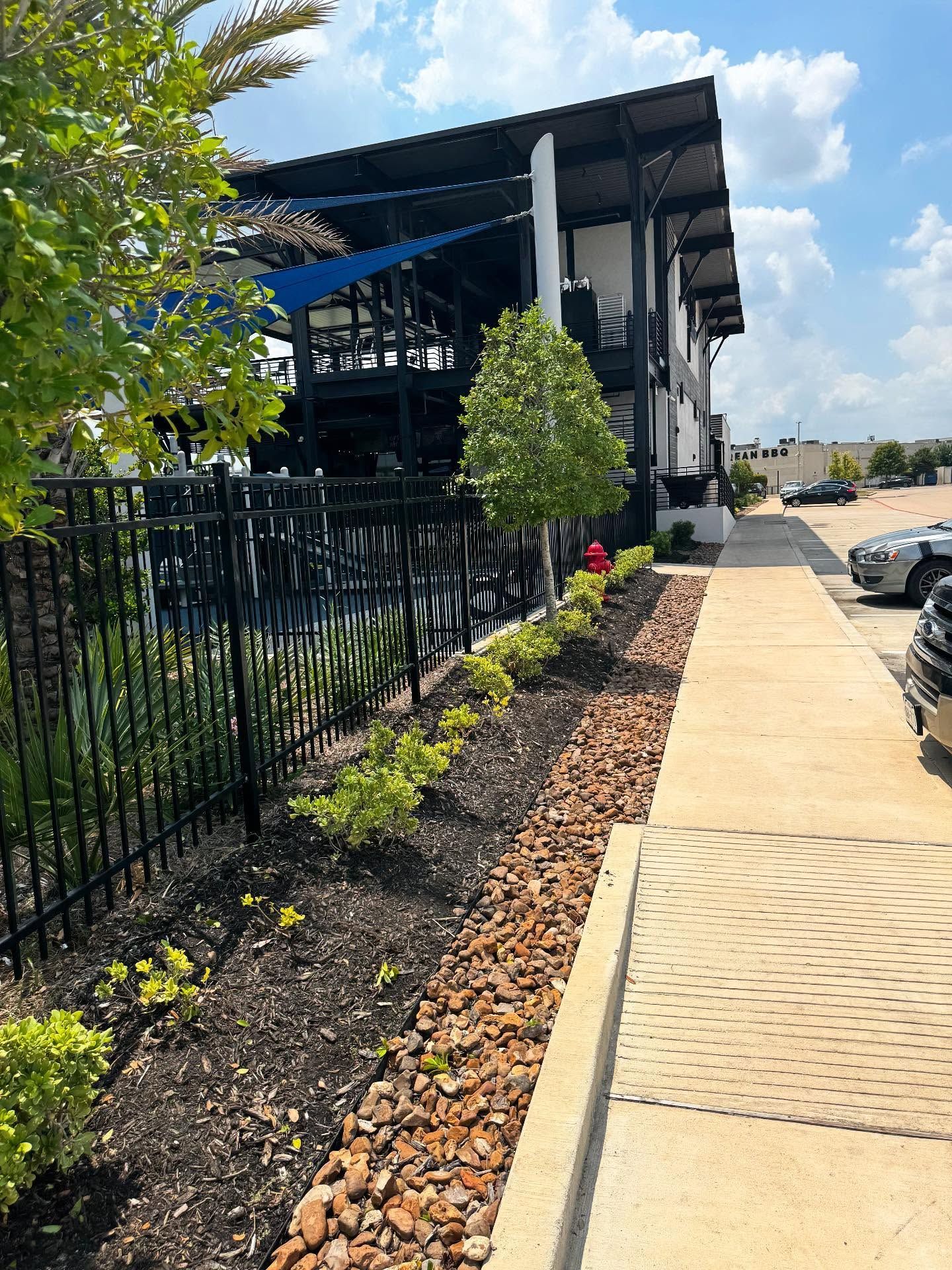 Exterior view of a two-story building with a black fence and sidewalk. Landscaping includes bushes, trees, and brown rocks.