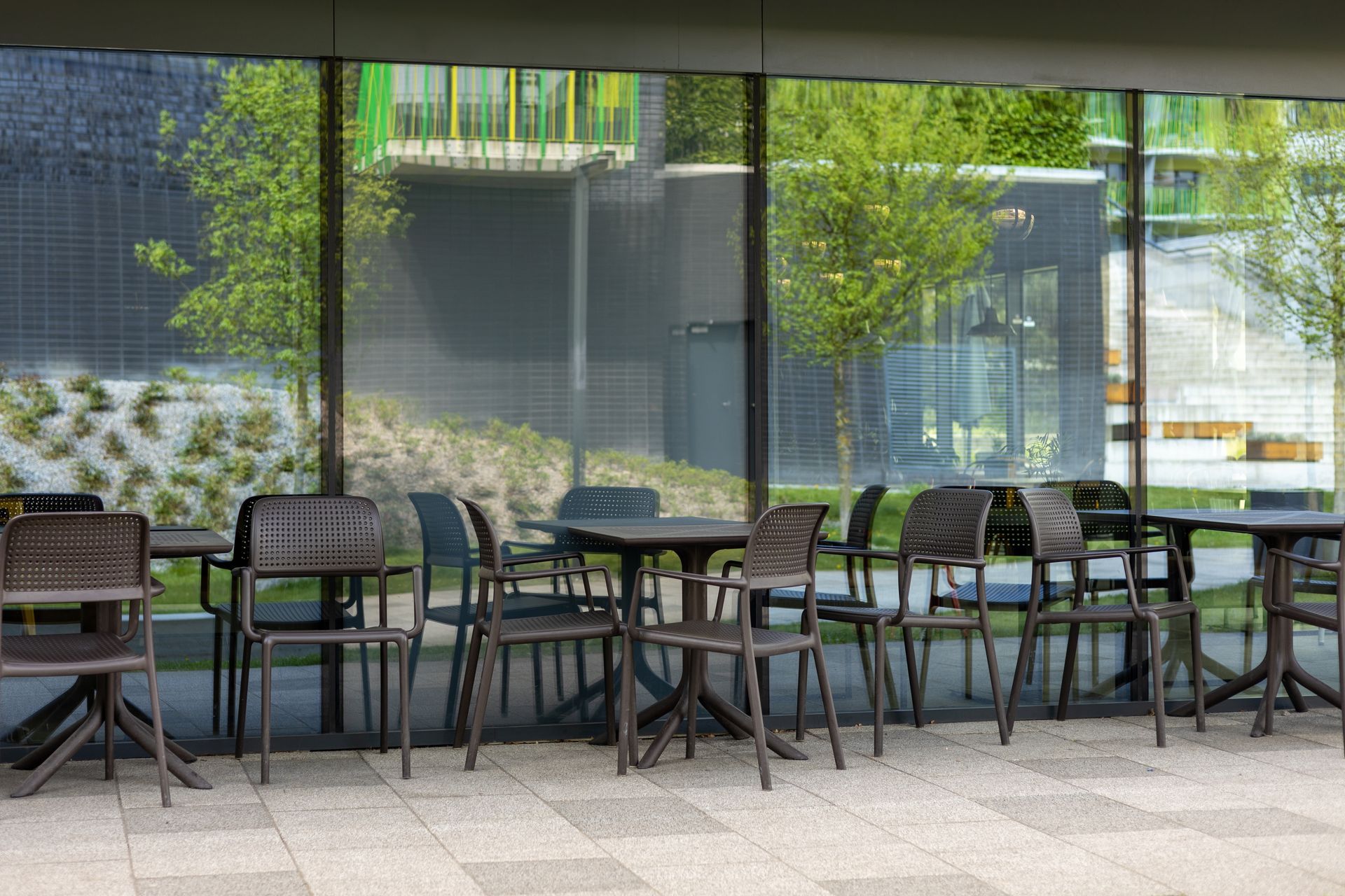 Outdoor seating area with tables and chairs in front of a glass building. Trees and green structures are visible.