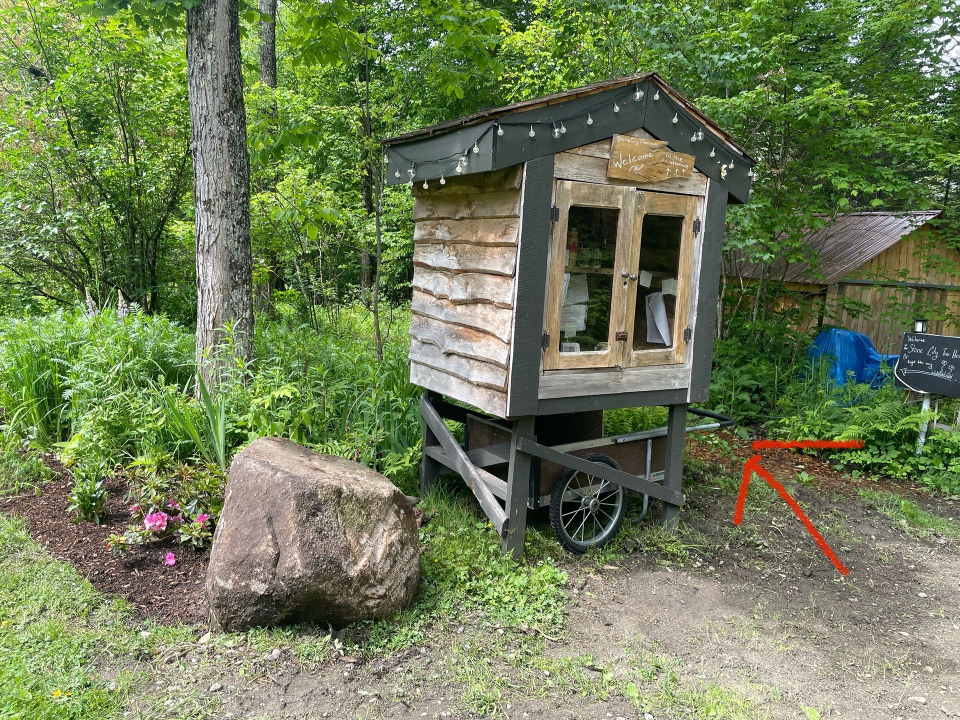 A small wooden cart with a miniature cabin on wheels, sits next to a large rock in a garden.