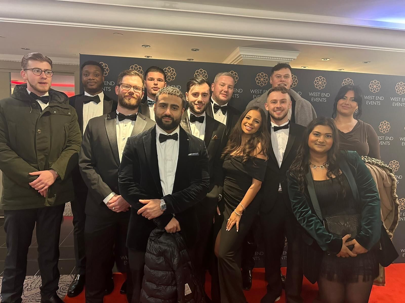 Group of people, mainly men in tuxedos, posing on a red carpet with a black backdrop.