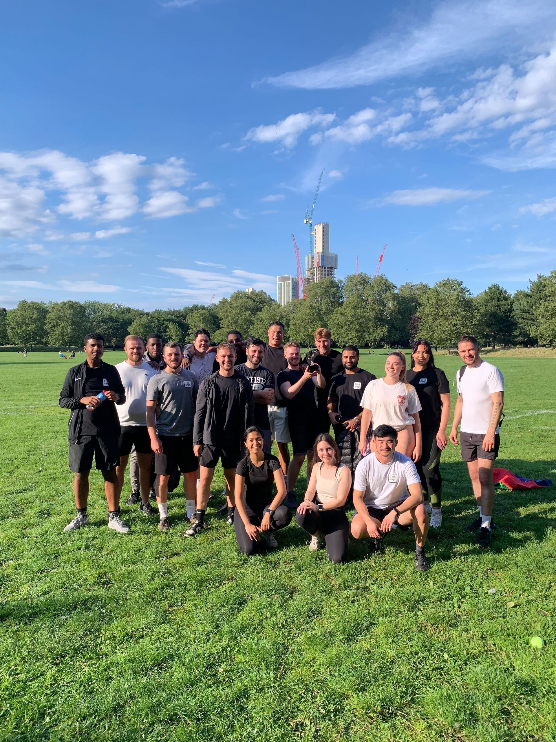 Group of people in a park, posing for a photo. Green grass, blue sky, and a distant building.