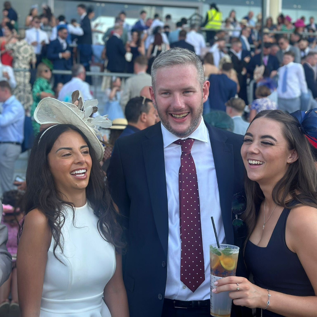 Three people smiling at a race track. A man in a suit is flanked by two women in dresses.