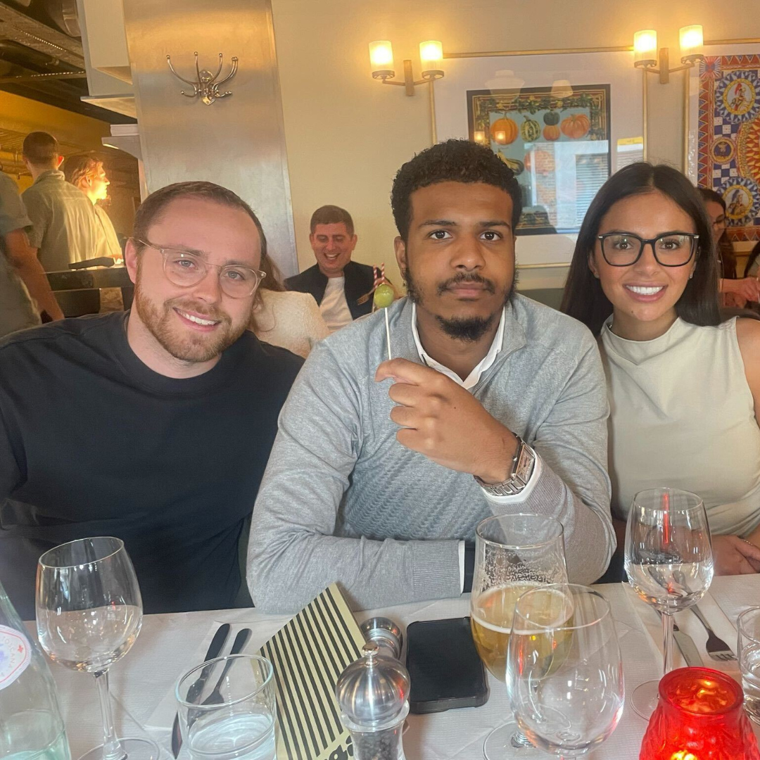 Three people at a table in a restaurant smiling, with wine glasses, a phone, and a lantern.