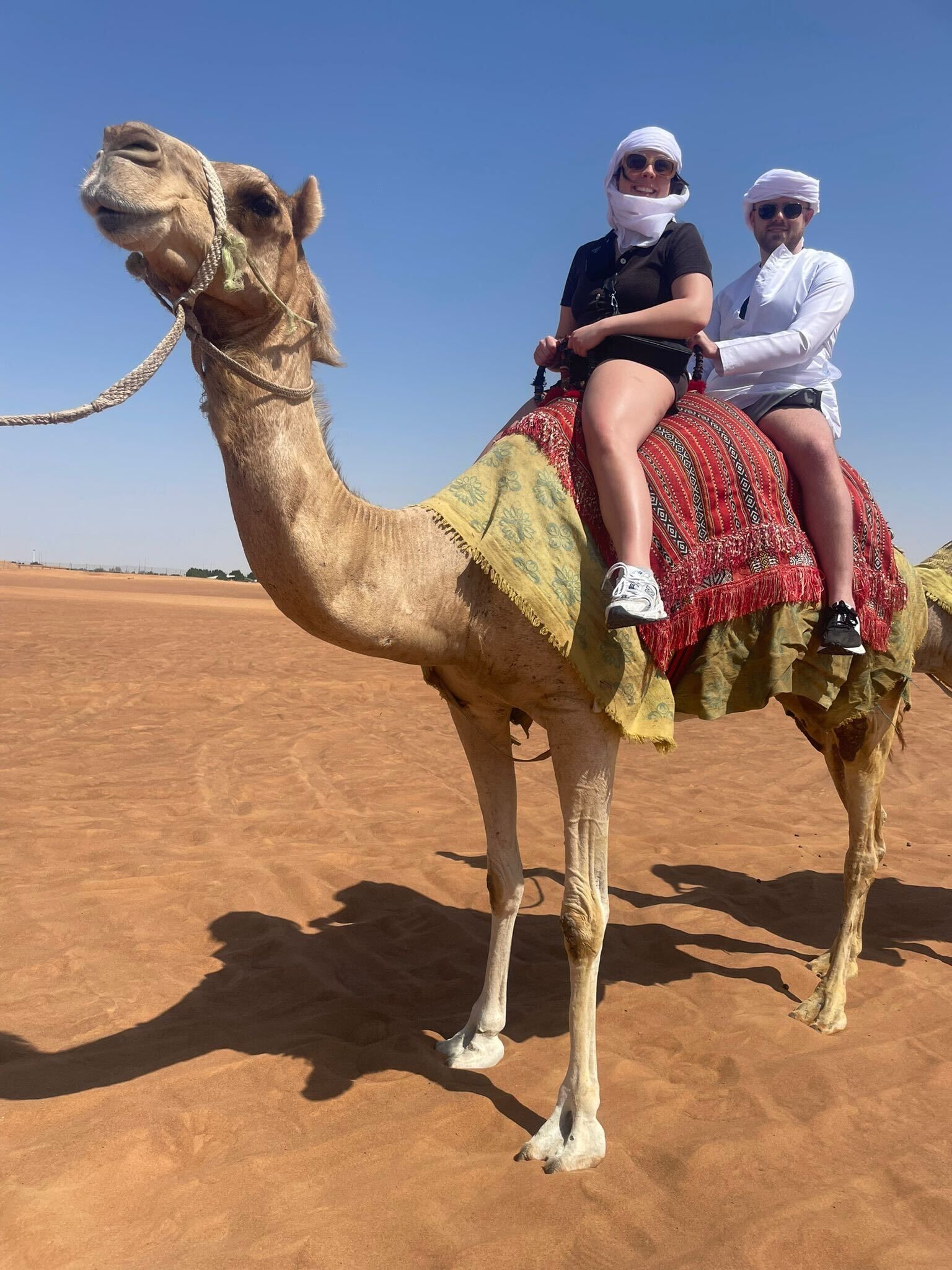 Two people riding a camel in a desert setting, under a clear blue sky.