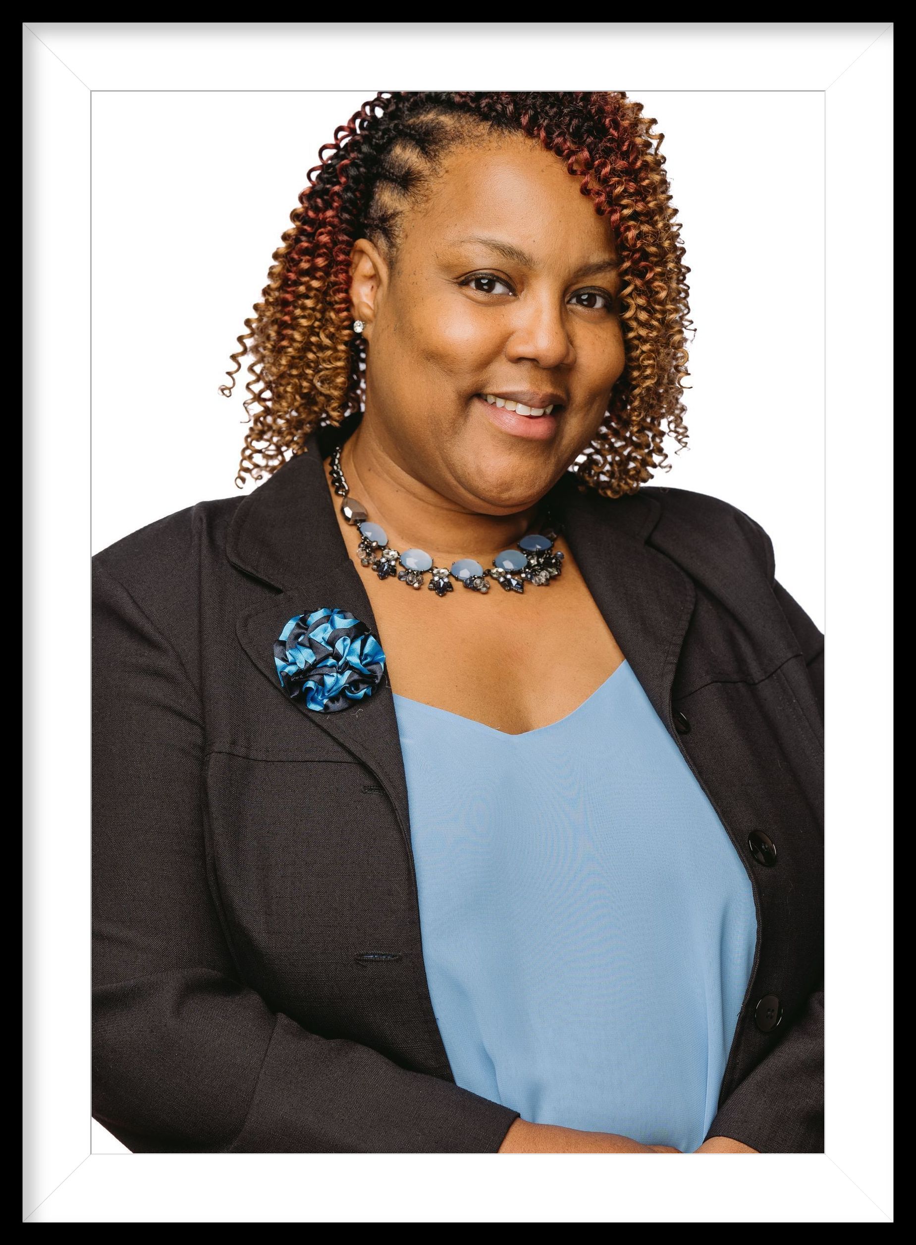 A woman is smiling, wearing a blue necklace, blue camisole, black blazer with a blue boutonniere