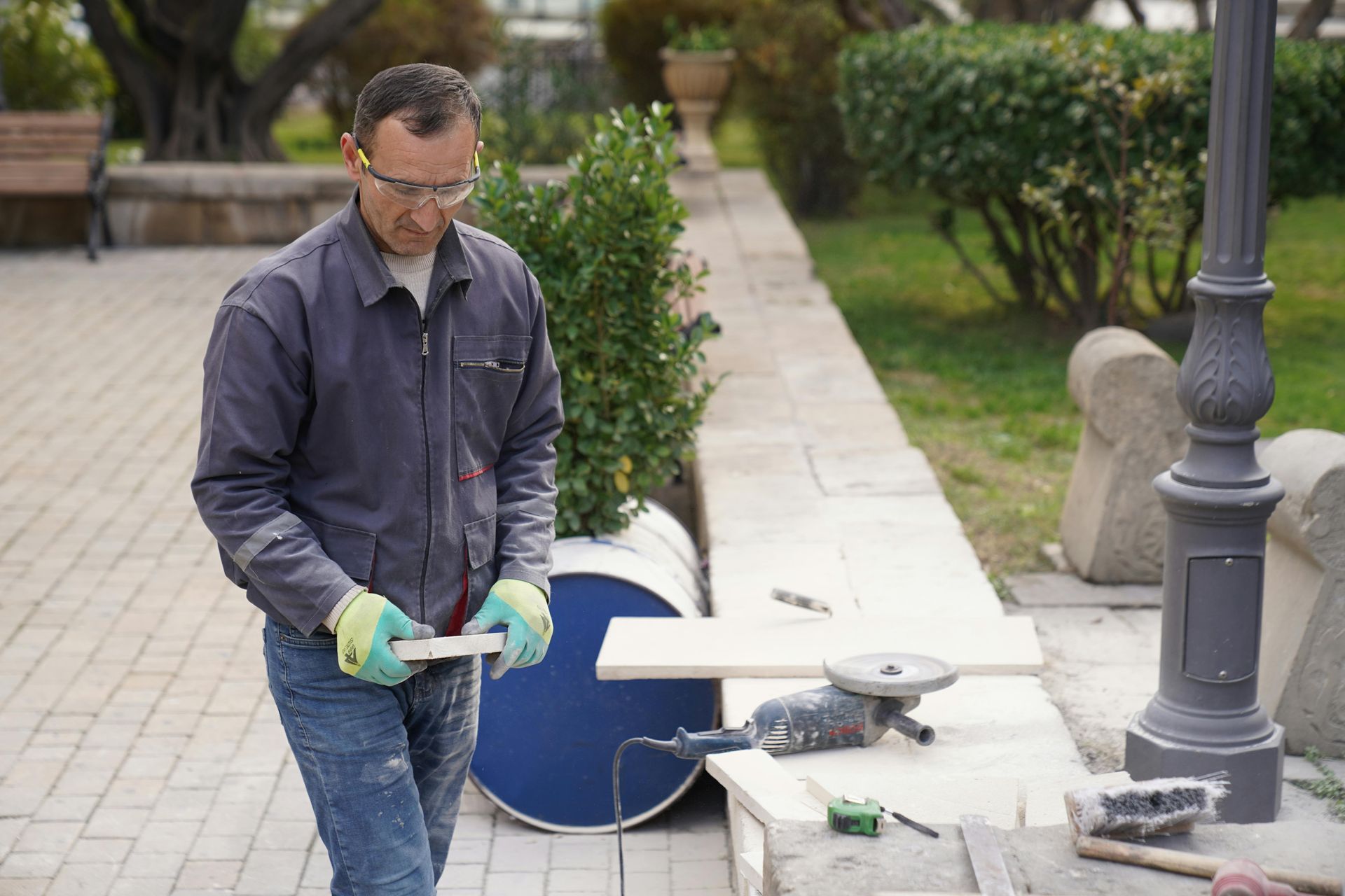 Man in safety glasses and gloves working with wood outdoors, tool nearby.