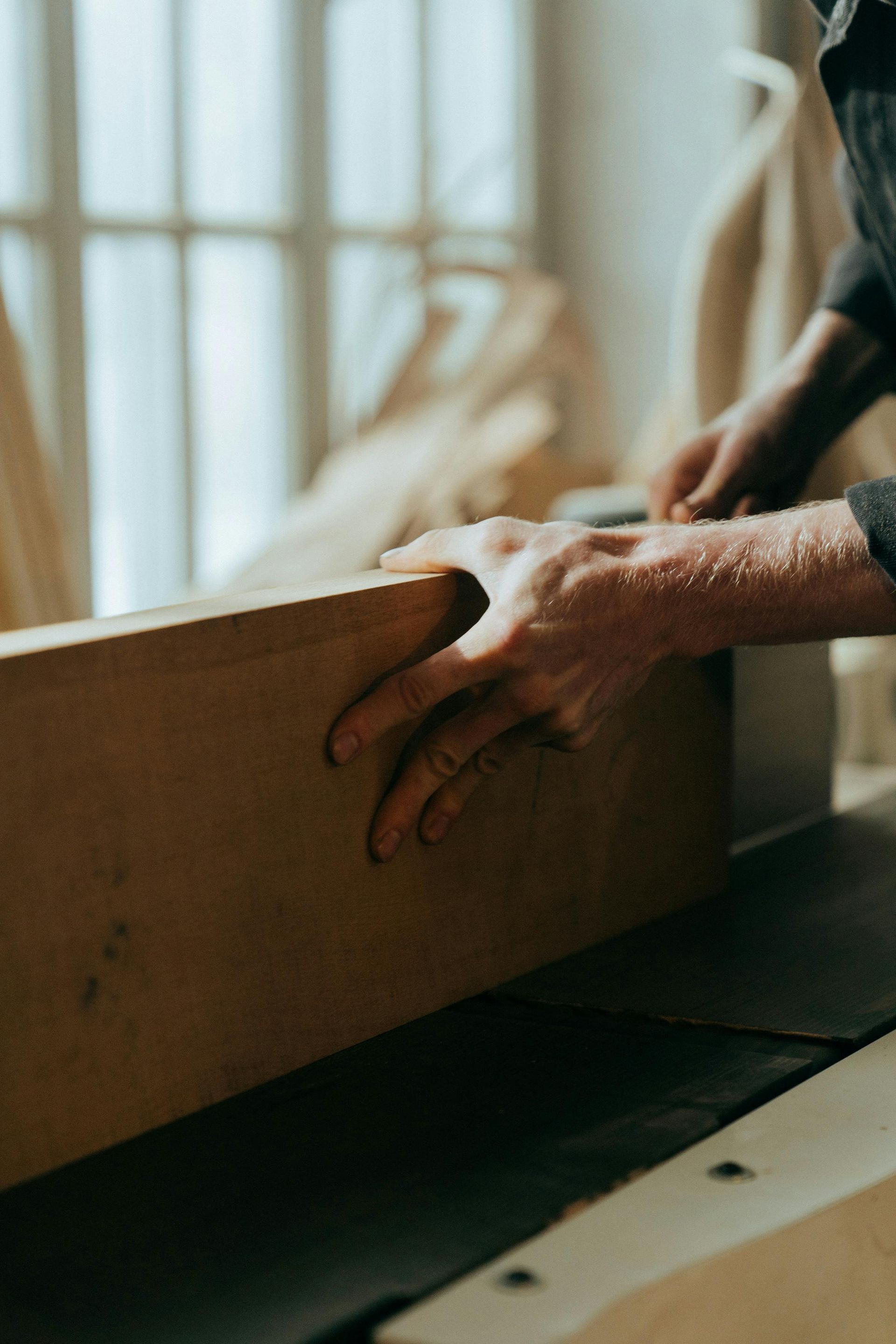 Hands holding a wooden plank, guiding it through a saw in a workshop.