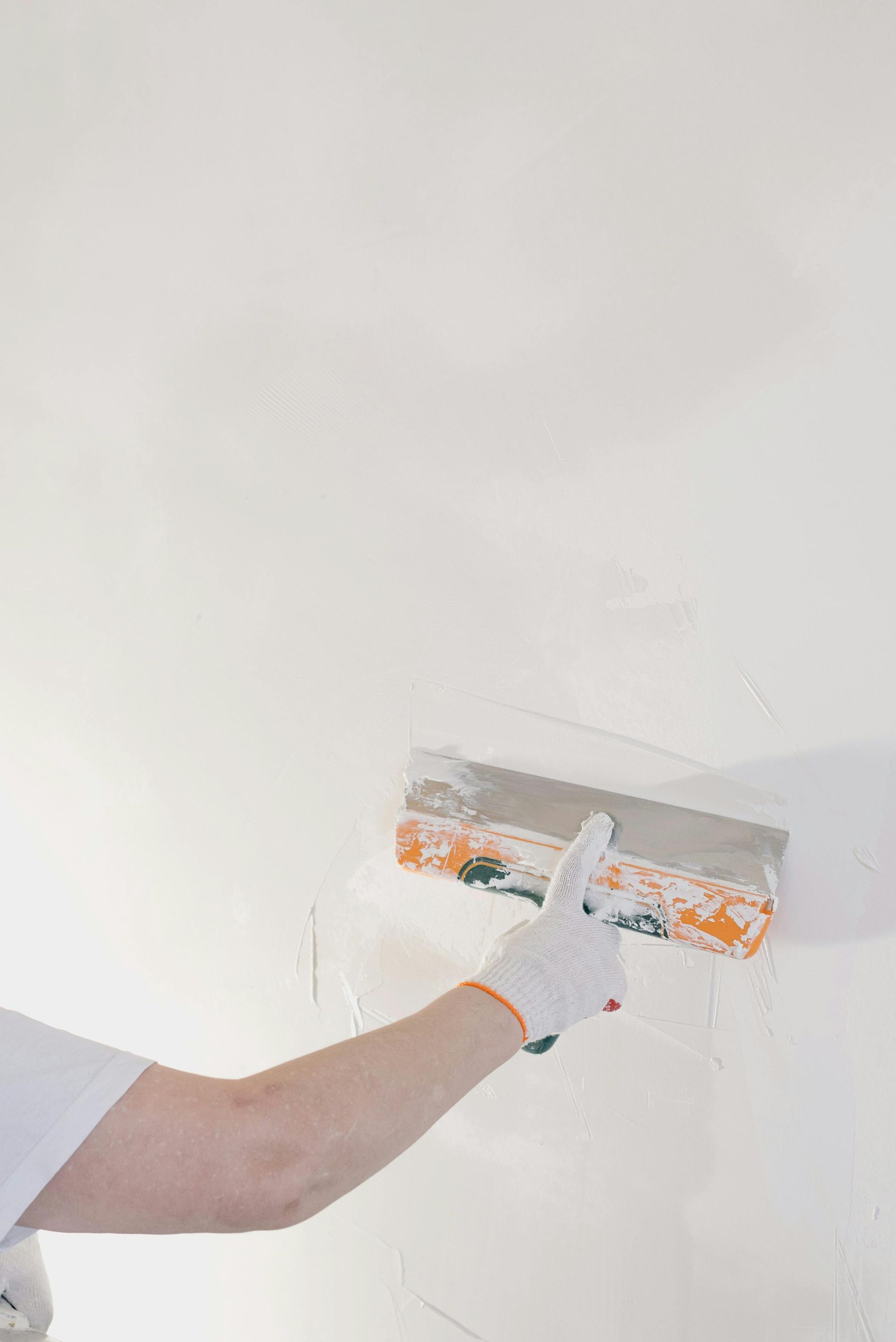 Person in a white glove uses a putty knife to smooth white plaster on a wall.