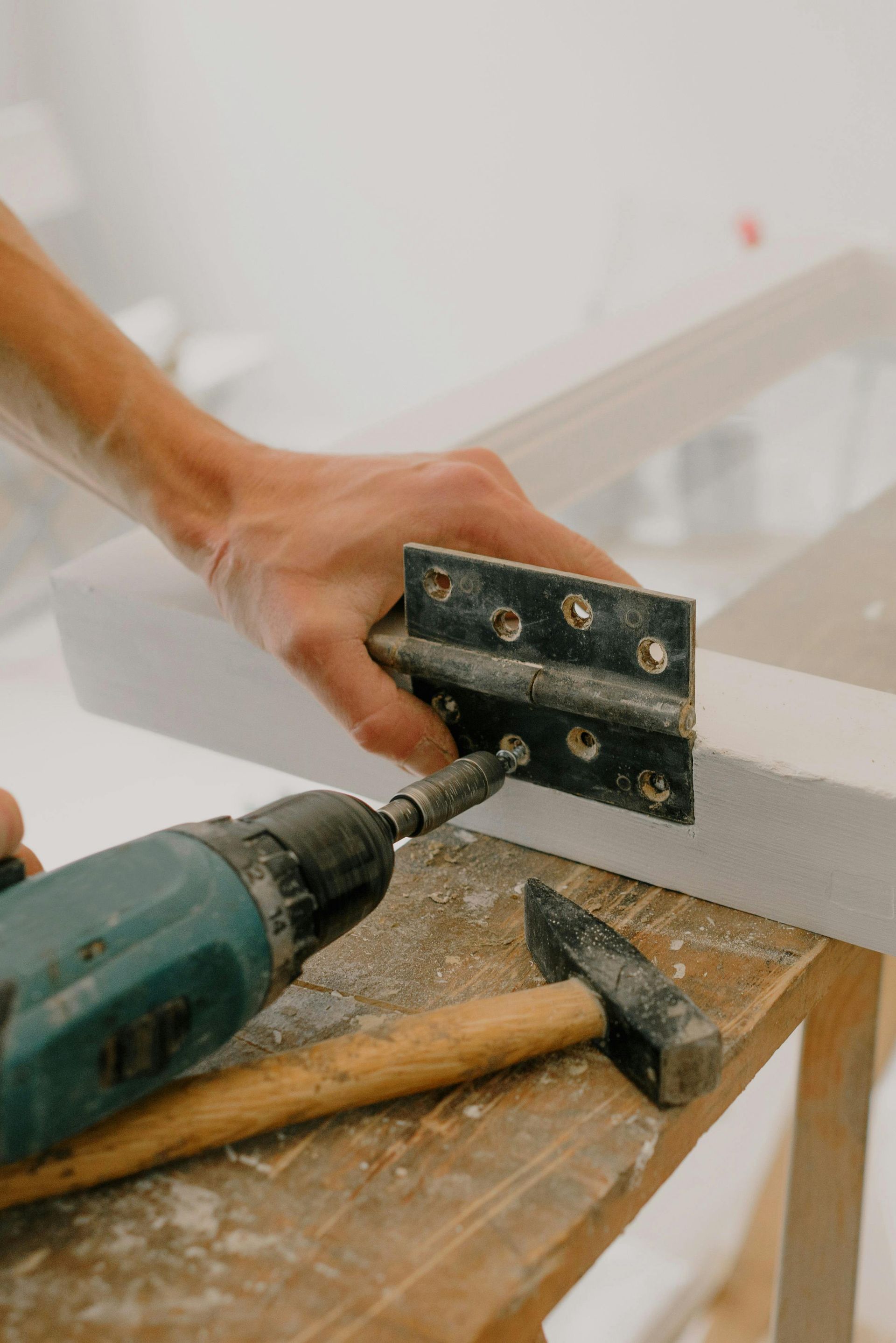 Person using a power drill to attach a hinge to a white wooden frame on a work table.