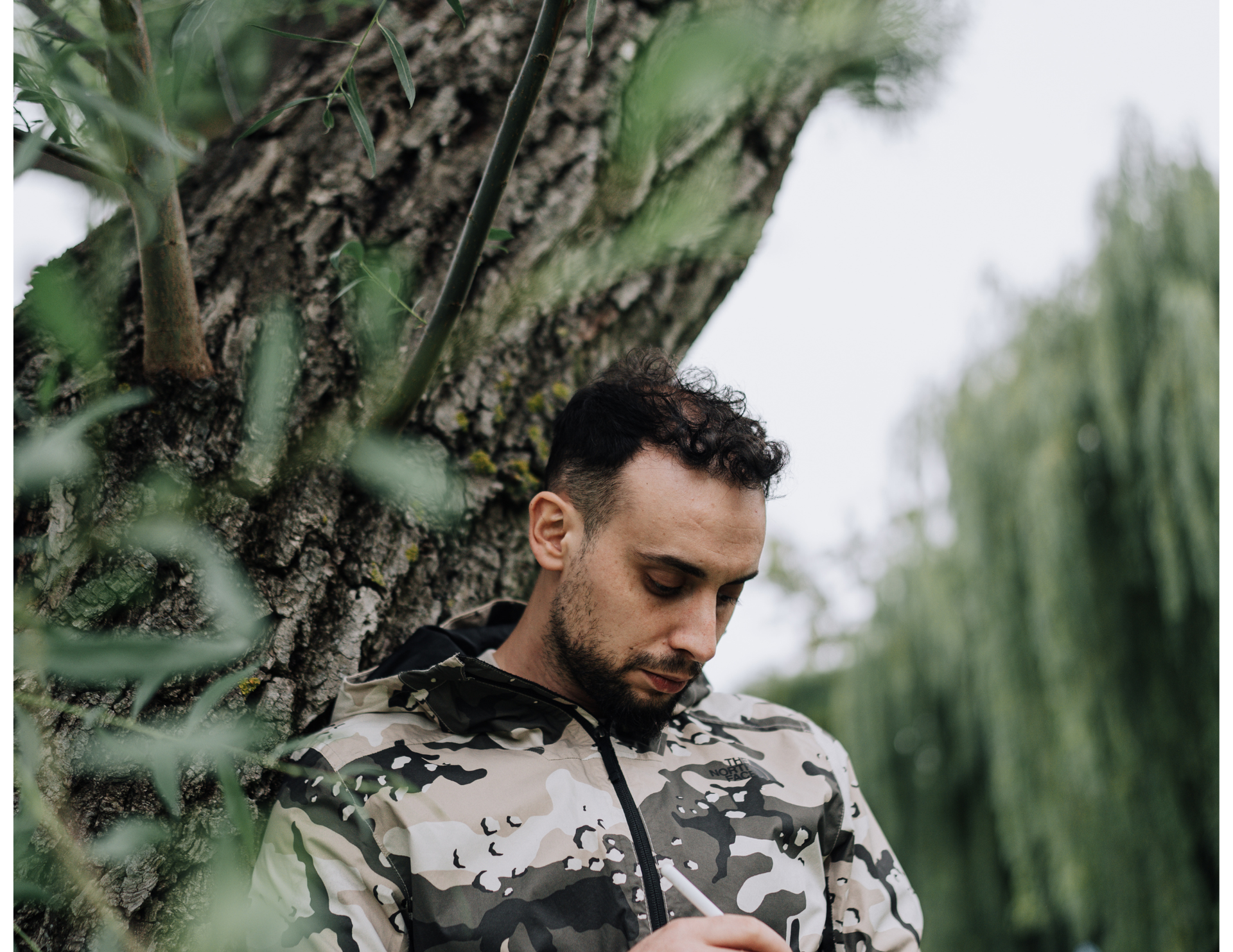 Man in camo jacket leaning against a tree, looking down, outdoors.
