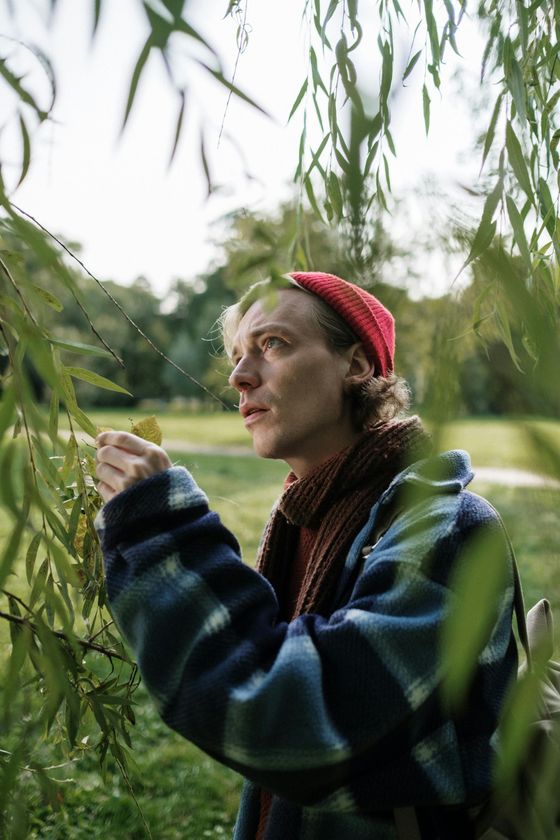 Man in red hat examines leaves under a willow tree, blue and brown patterned jacket, outdoor setting.