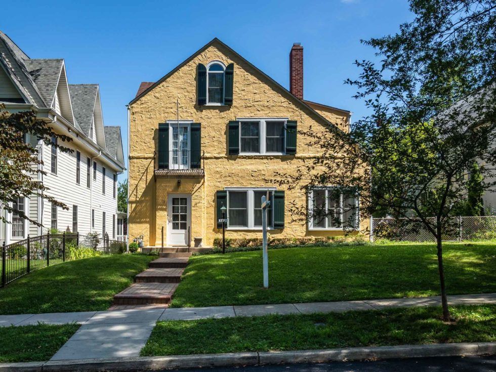 Yellow house with green shutters, brick path, and a grassy yard on a sunny day.