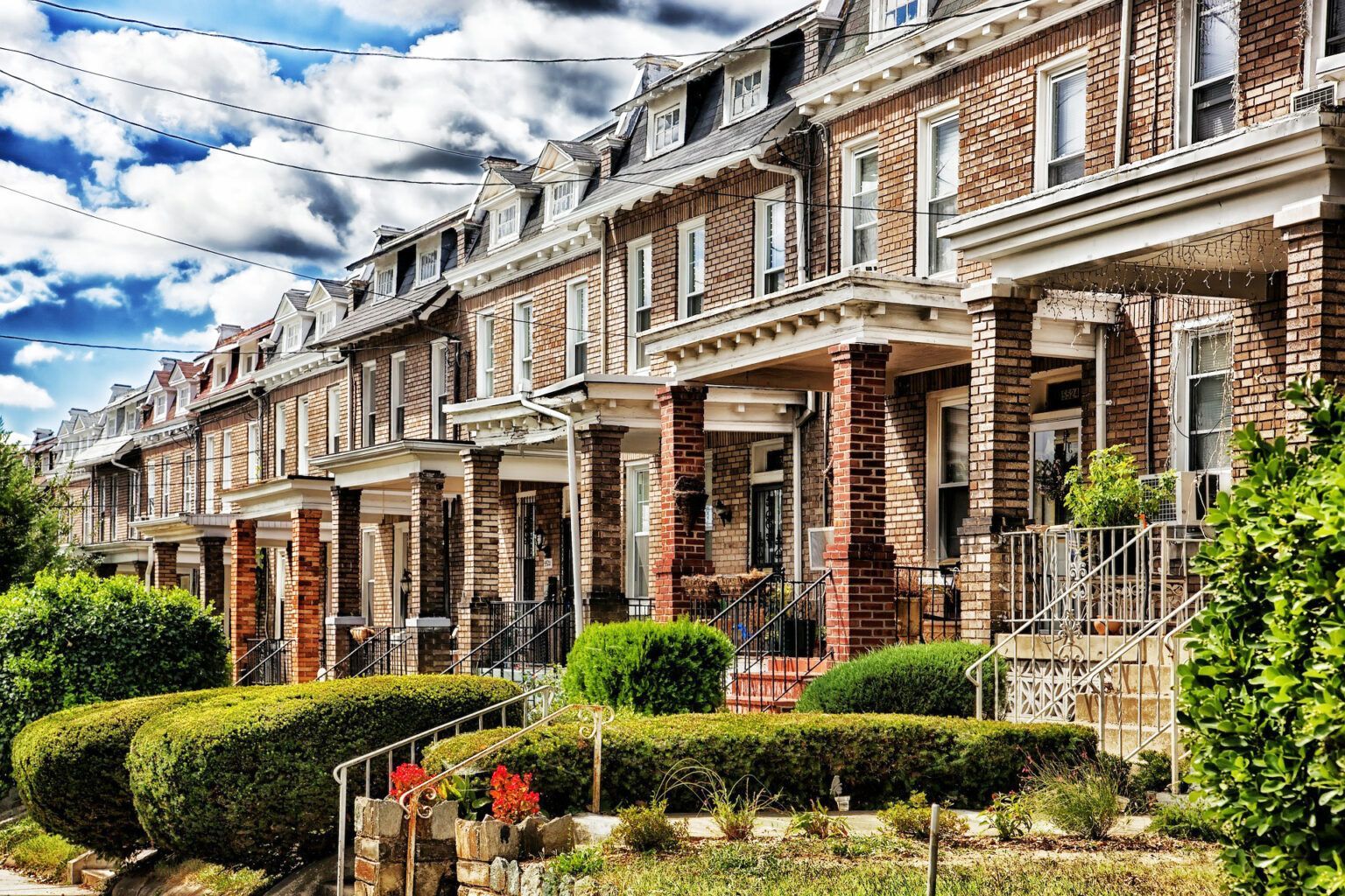 Row of brick rowhouses with porches under a blue sky with clouds. Lush green bushes line the front yards.