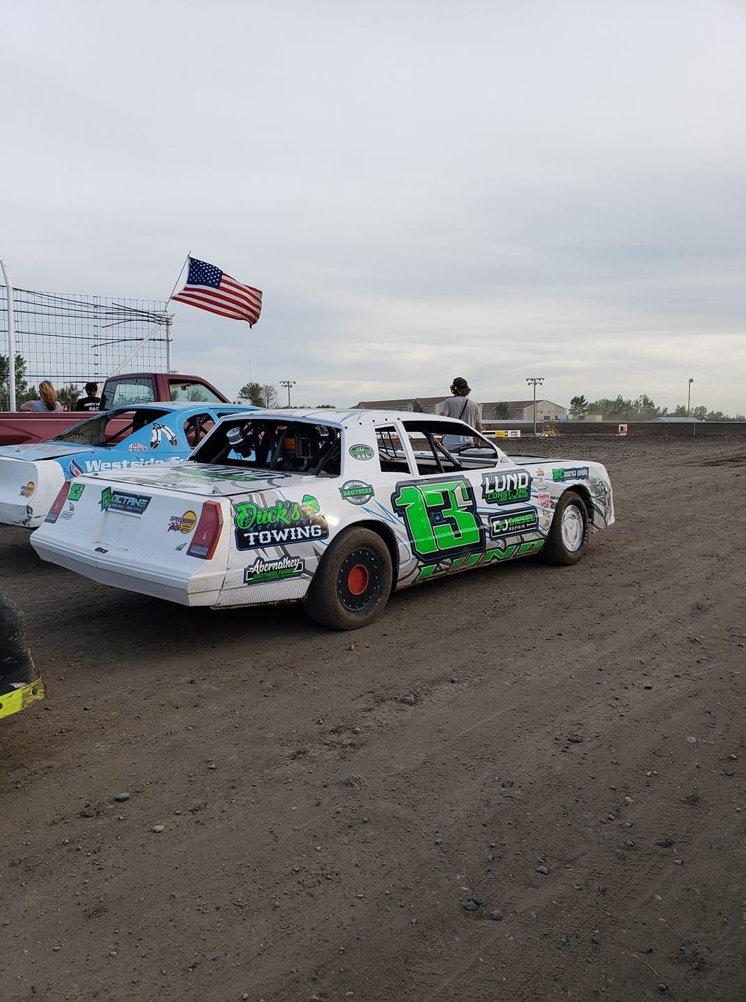 White rally car with sponsor decals on a dirt track, waving an American flag under a cloudy sky.