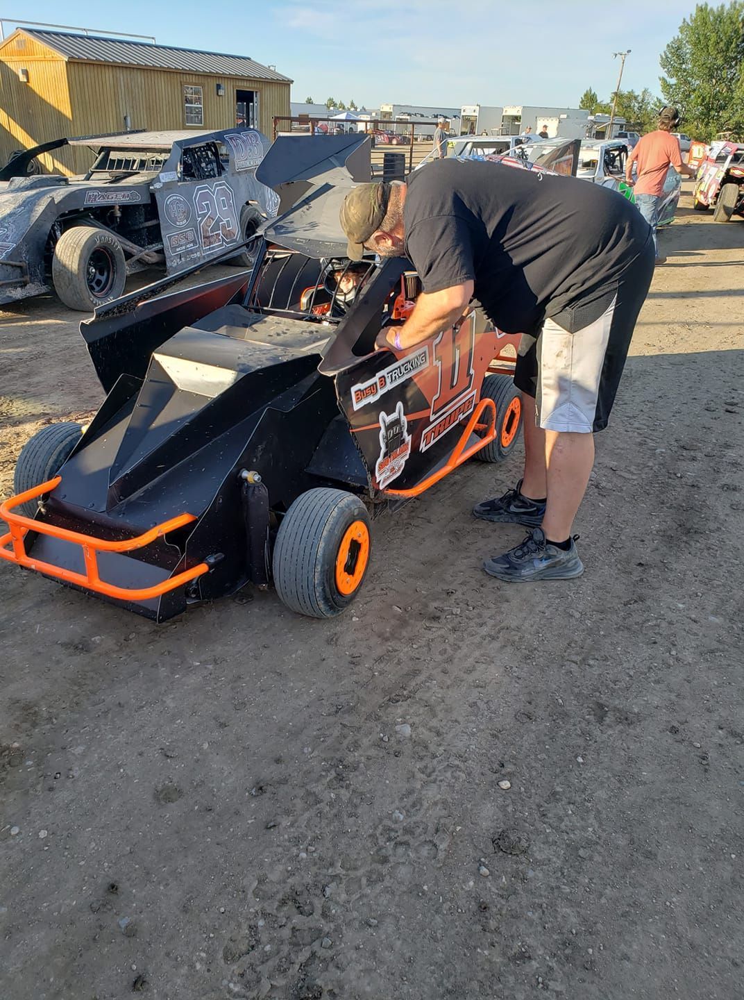 Man bent over a black-and-orange go-kart in a dirt lot, working on it beside other parked carts.