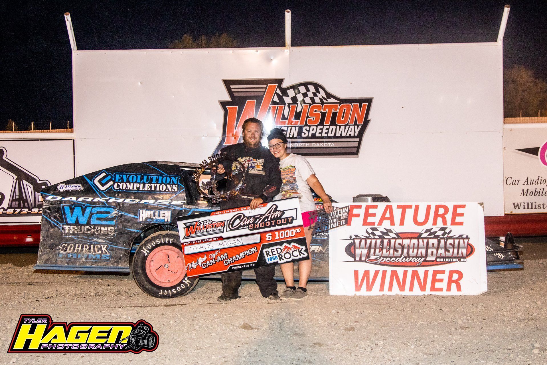 Race car driver posing with a black-and-orange dirt car beside a “Feature Winner” sign on a track.