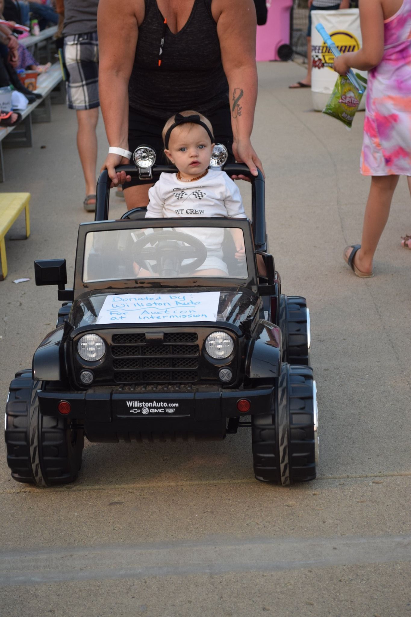 Toddler riding in a black toy jeep while an adult pushes from behind on a crowded walkway