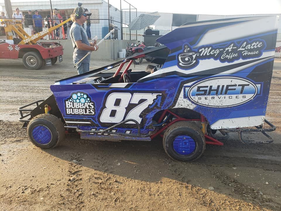 Blue and black number 87 race car with sponsor logos, parked on dirt at a dirt track with a driver beside it.