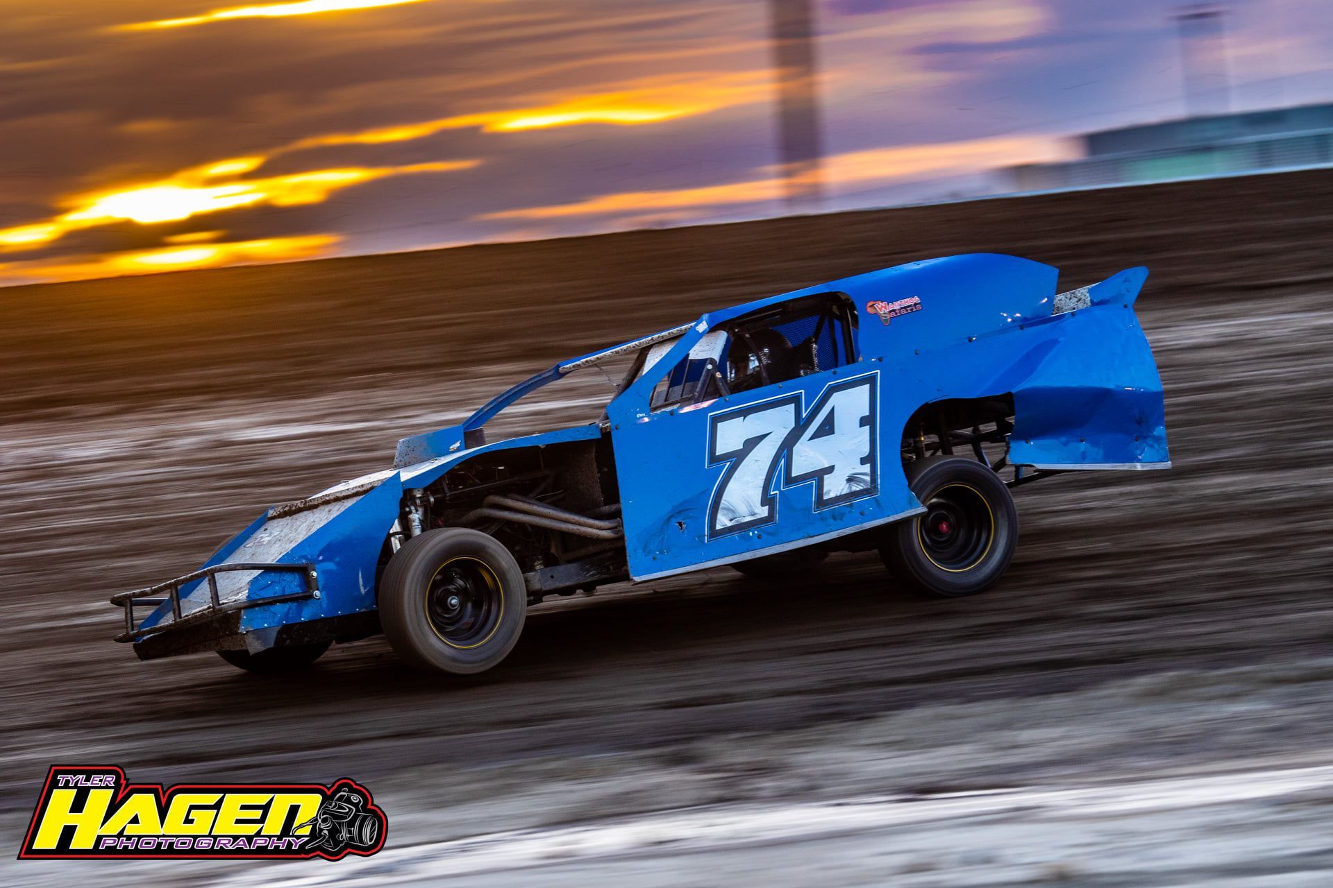 Blue #74 race car drifting on a dirt track at sunset