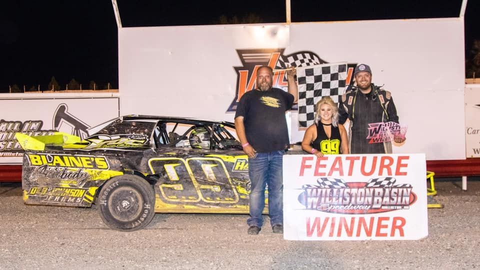Racetrack winner poses with car and checkered flag; two people hold a “FEATURE WINNER” sign.