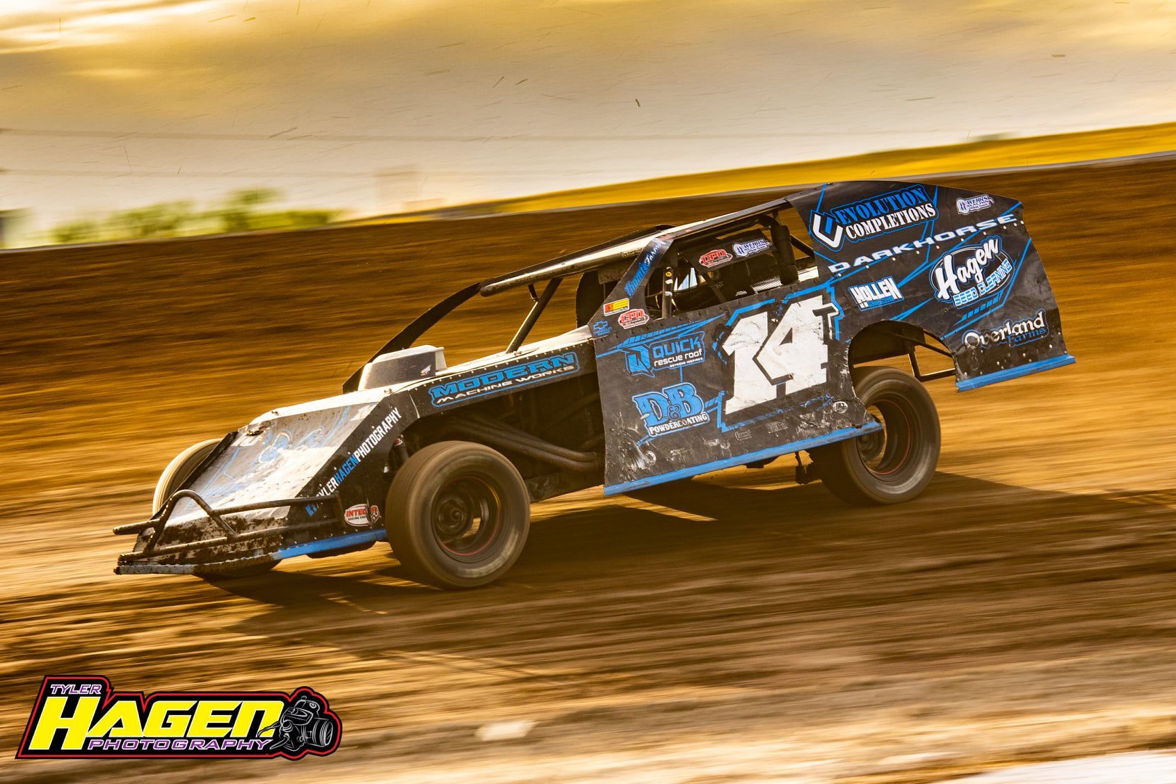 Blue dirt race car sliding on a dirt track at sunset, with sponsor decals and number 14 on the side