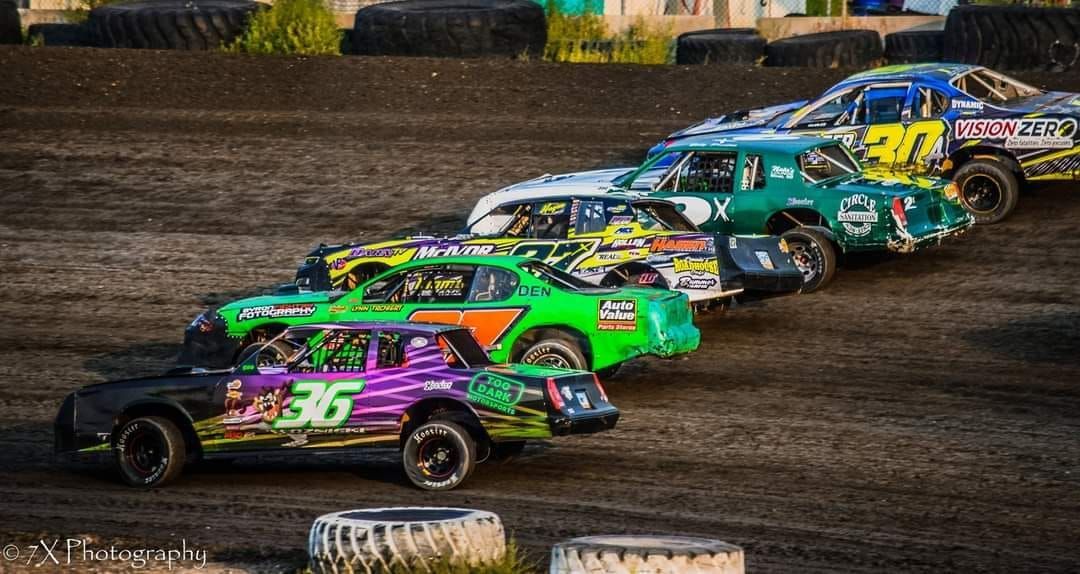 Stock cars racing side by side on a dirt track, with a purple and green car leading the pack