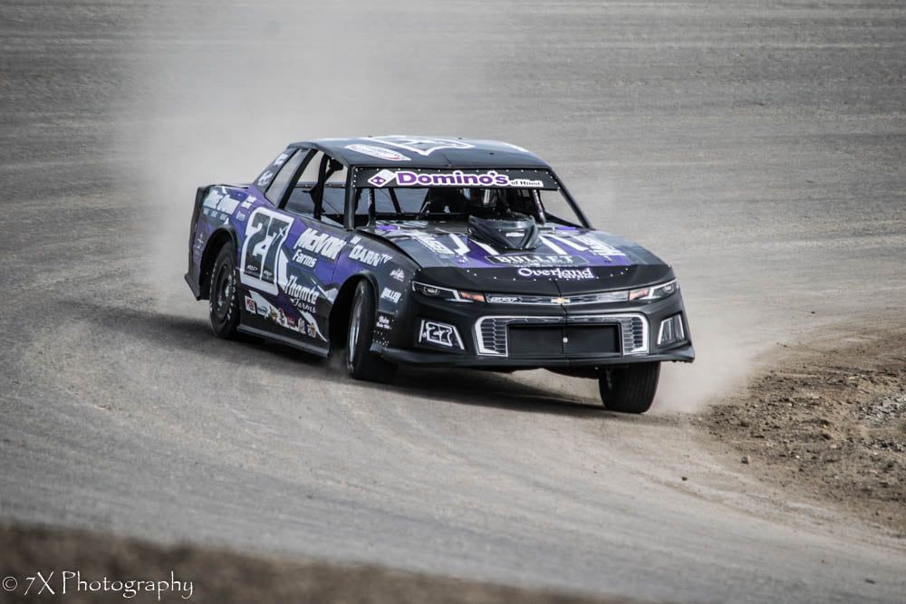 Blue stock car drifting on a dirt track, kicking up dust around a sharp turn.