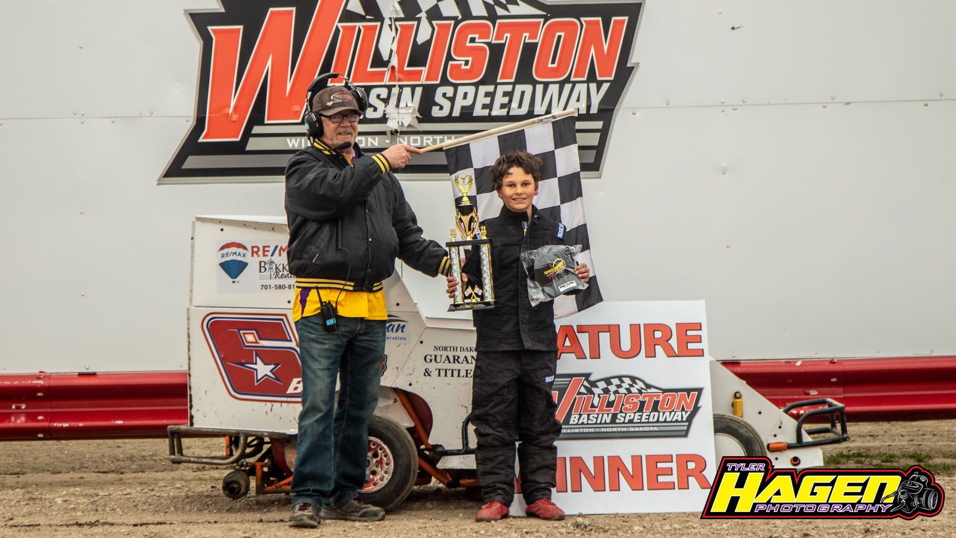 Two race drivers holding trophies on a dirt track podium at Williston Basin Speedway, with winner banner behind them