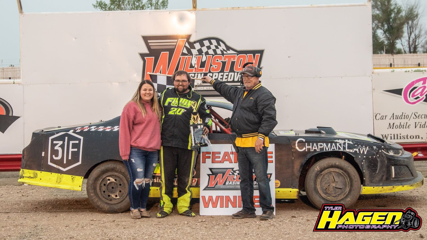 Three racecar drivers posing in front of a dirt track car with sponsor logos and a victory backdrop.