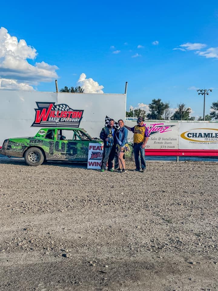 Four people posing beside a green and white drag racing truck on a gravel lot under a blue sky.