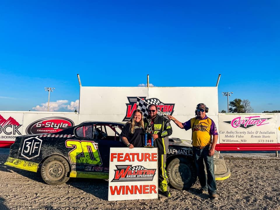 Race car team posing with a “Feature Winner” sign beside a black and yellow dirt track car under a clear blue sky.