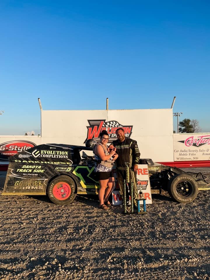 Race car and driver standing in front of a black dirt track car at sunset, with trophies on the ground.