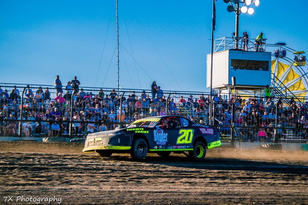 Race car number 20 speeding on a dirt track with a crowd in the stands behind it.