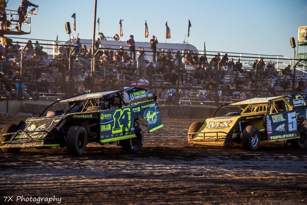 Two dirt track race cars racing side by side at dusk with a crowd in the stands.